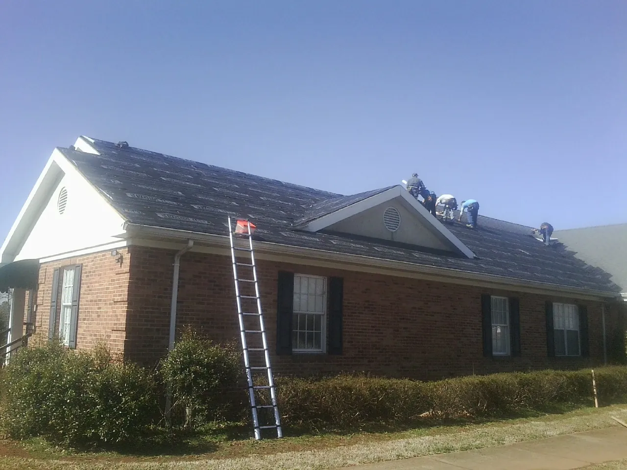 Crew tearing shingles off a brick ranch roof with a ladder set — Emergency Roof Repair work by our crew in Ida