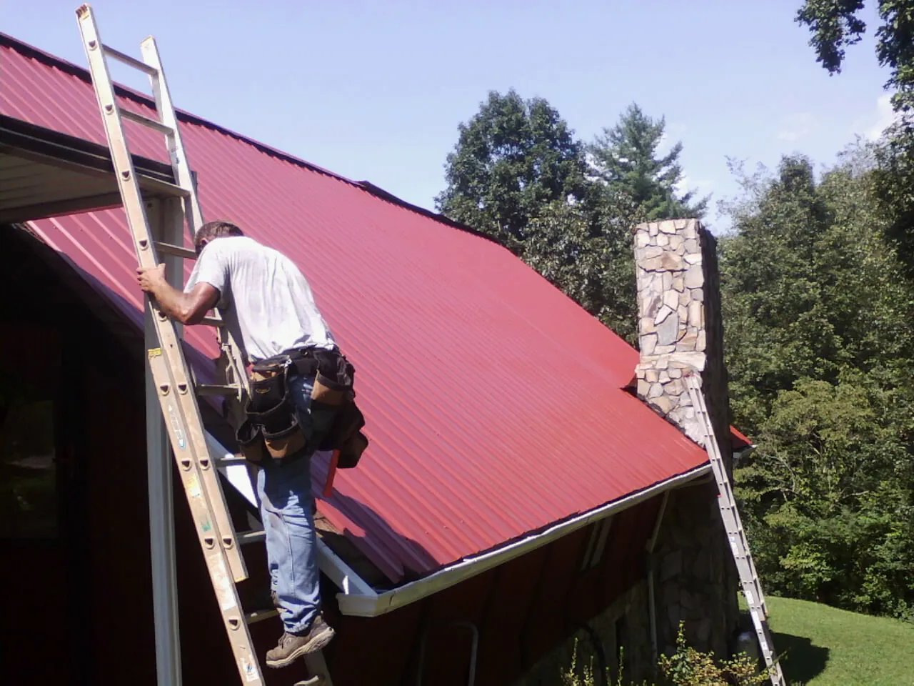 Roofer on a ladder beside a red metal roof and stone chimney — Soffit & Fascia Repair by Roofing Brothers in Ida